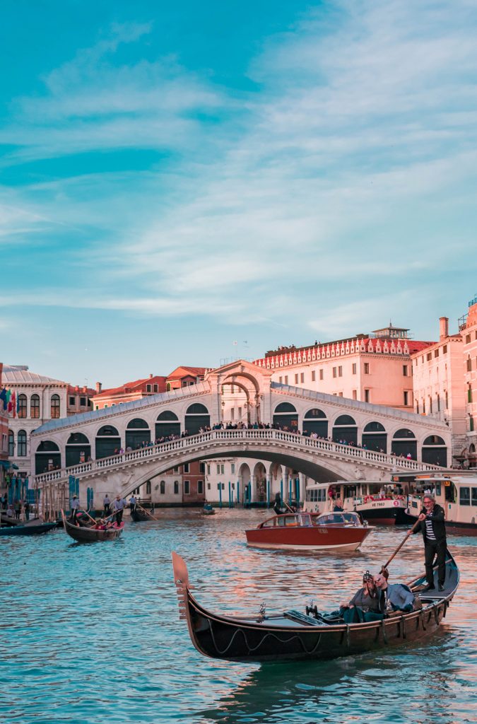 Eine Gondola mit einem Gondoliere und einem Paar vor einer Brück in Venedig. 