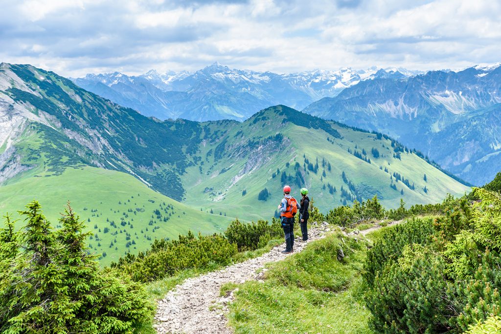 Wanderer in grüner Berglandschaft. Im Hintergrund sind schneebedeckte Gipfelketten zu sehen.