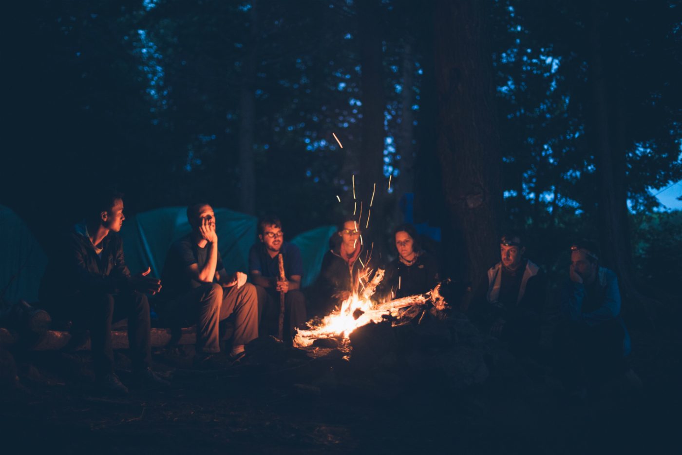 Eine Gruppe von Menschen sitzt um ein Lagerfeuer. Im Hintergrund sind Zelte zu sehen.