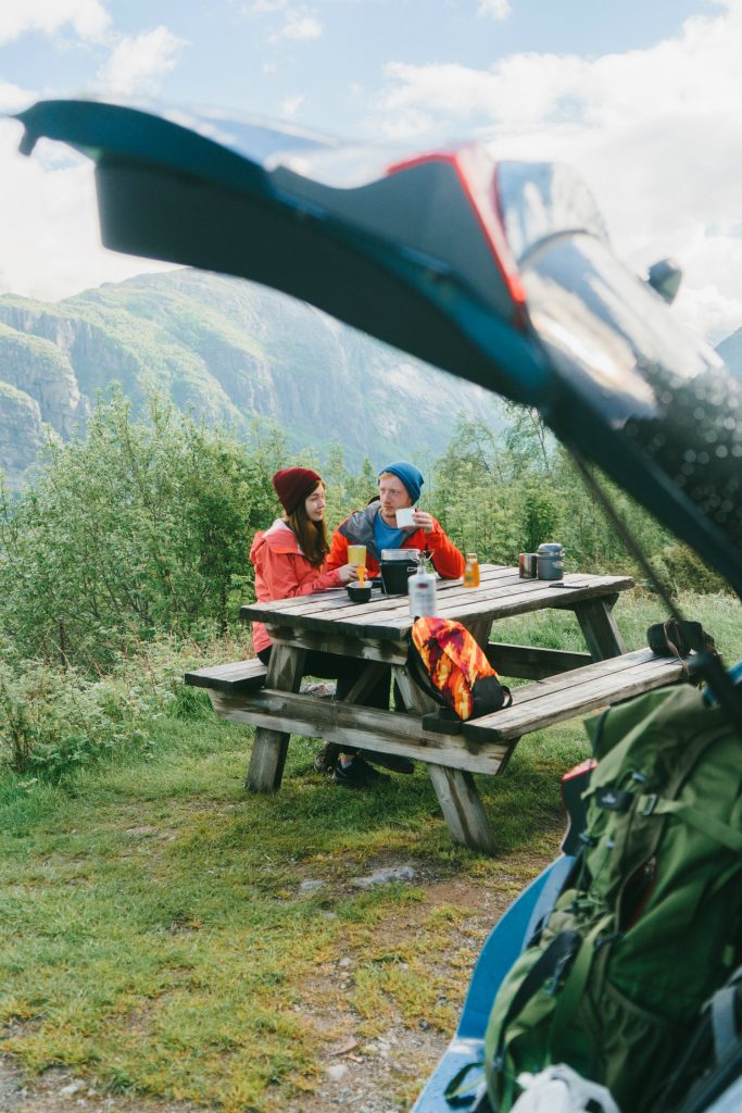 Paar sitzt an einem Tisch im Freien. Im Vordergrund ist ein Kofferraum, im Hintergrund sind Berge zu sehen