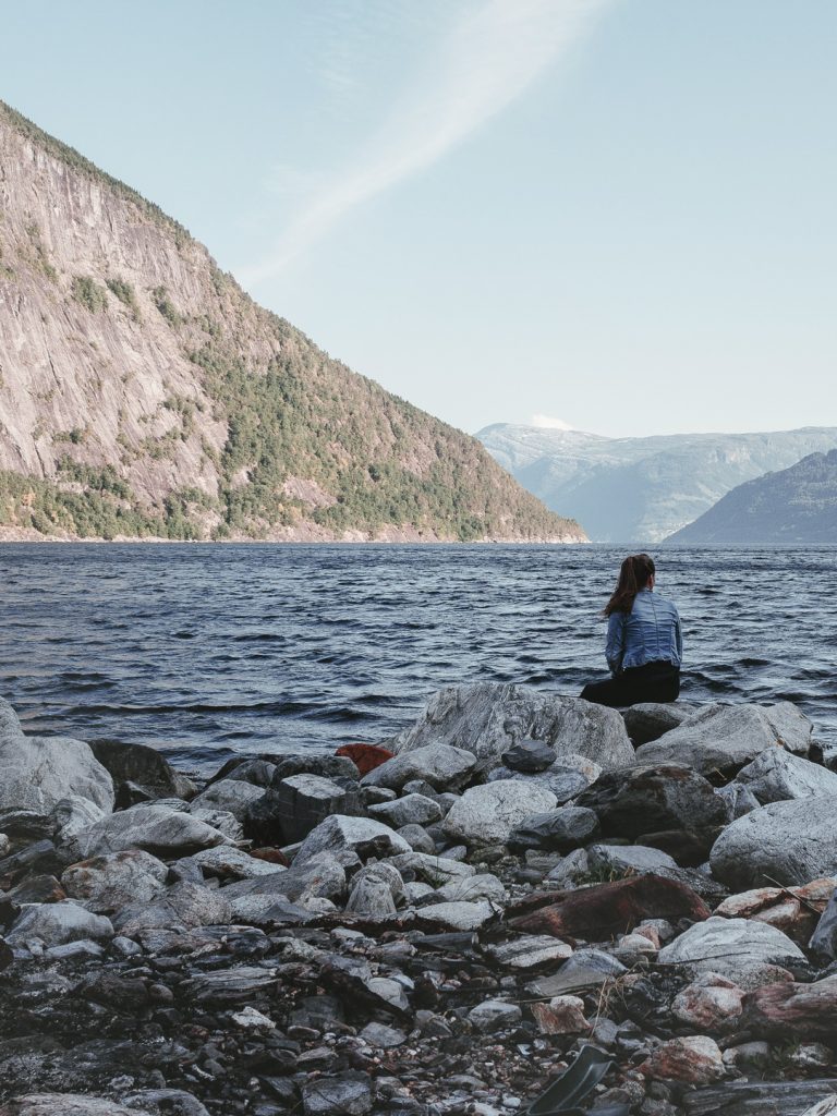 Stoppe mit dem Wohnmobil an einem der wunderschönen Fjorde Norwegens und lass die Natur auf dich wirken. Ein Frau sitzt am Ufer eines Fjordes in Norwegen.